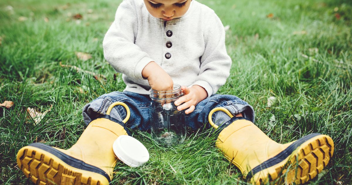 A boy sitting down on grass wearing yellow gum boots. He's opening a jar he's using to catch bugs.